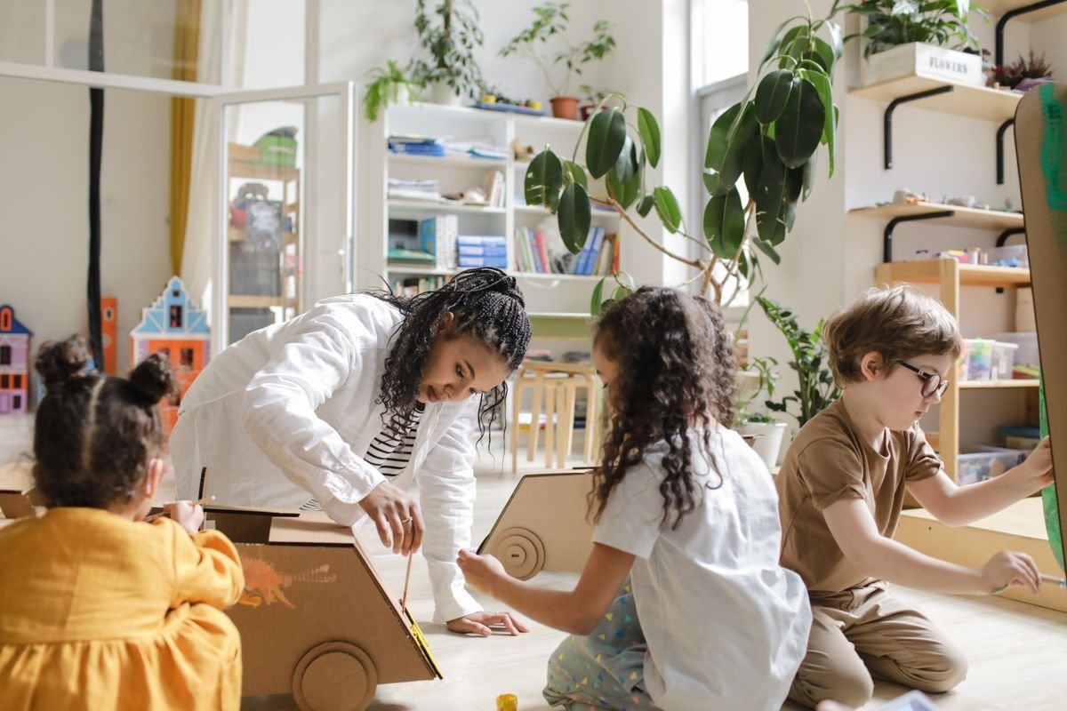 A teacher and children building a large cardboard model together in a bright, plant-filled room