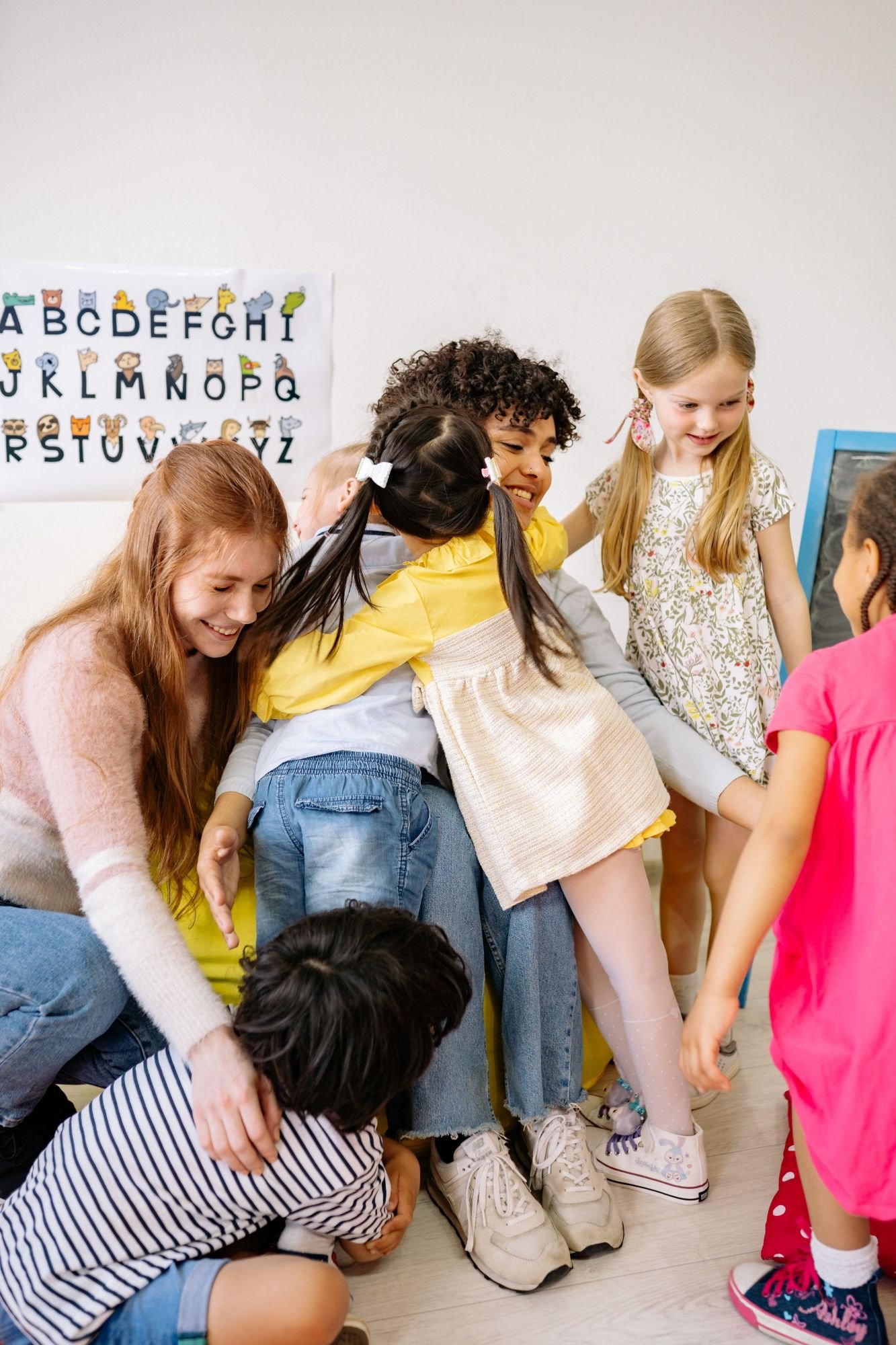 Two smiling teachers kneel on the classroom floor as a group of young children hug them