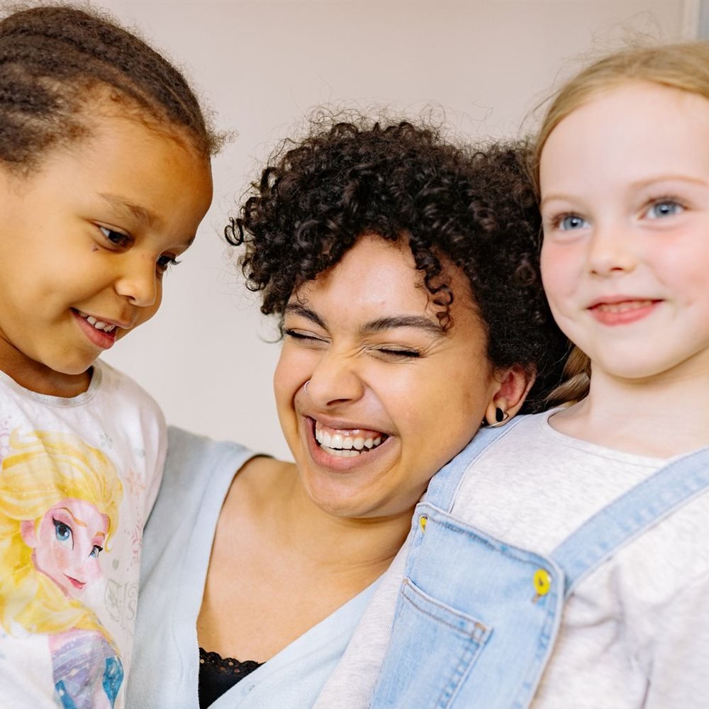 A smiling teacher with her arms around two young children in a classroom