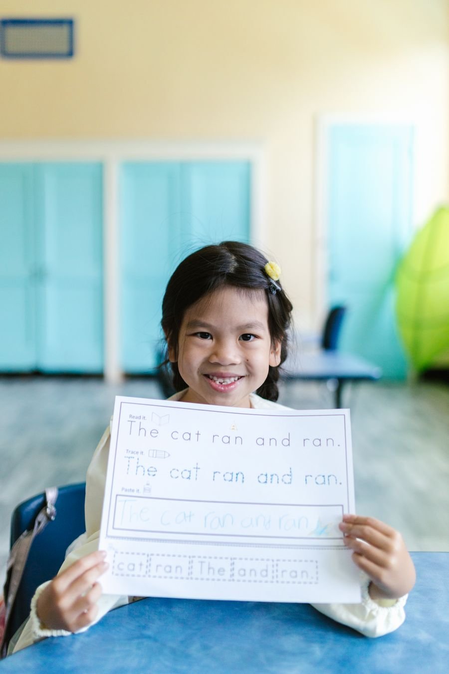 A young girl smiling as she holds up a writing worksheet she has finished