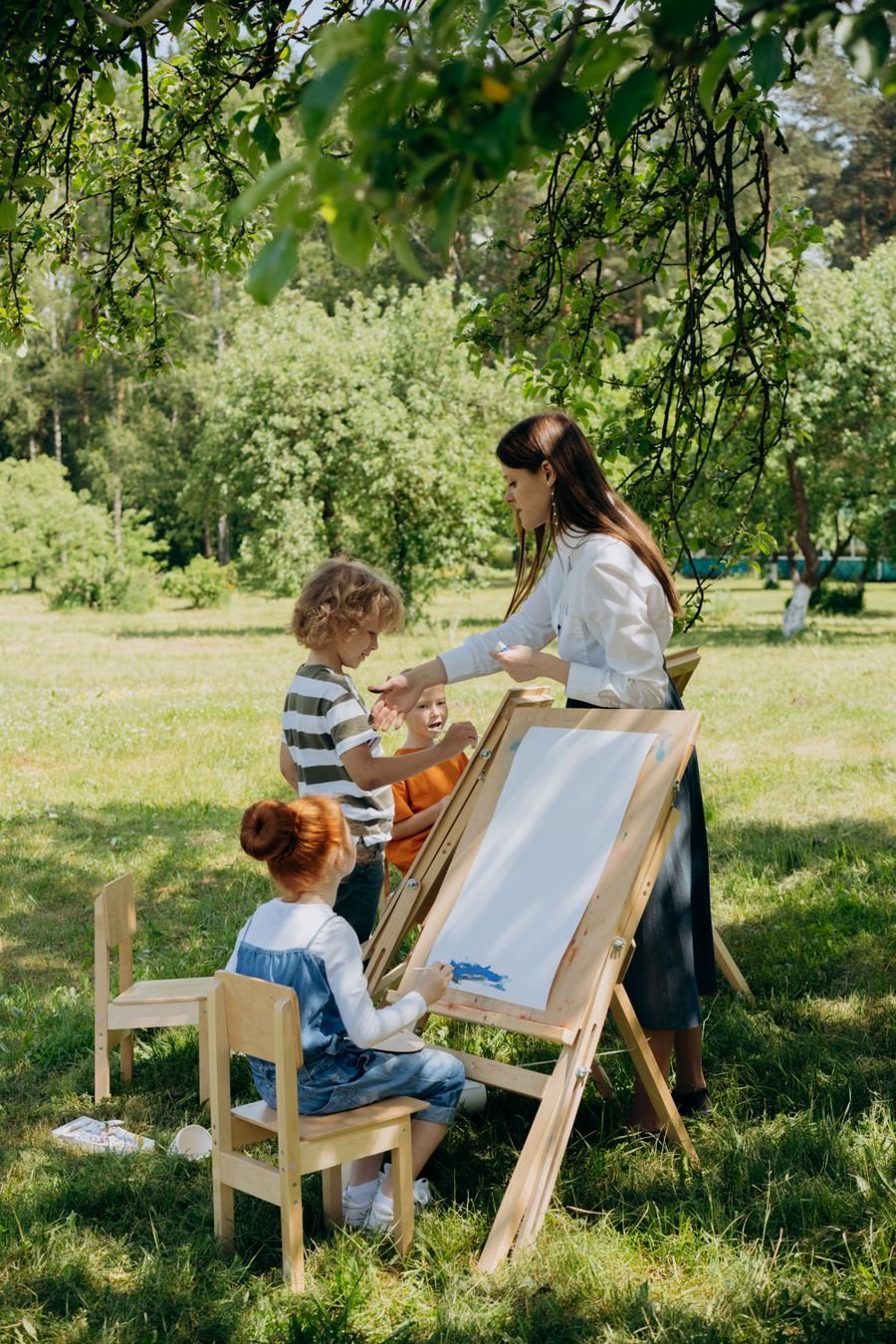 A teacher and three children painting at easels on the grass under trees