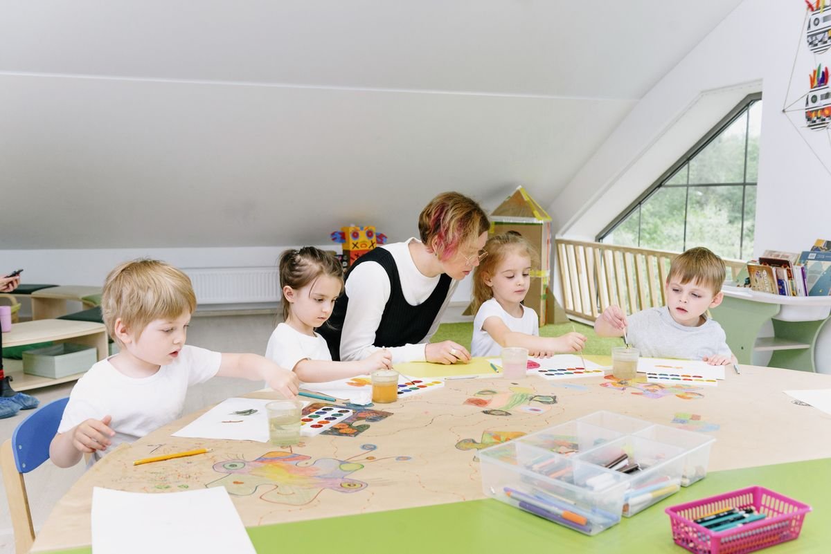 Children working at a table with art materials while a teacher guides them