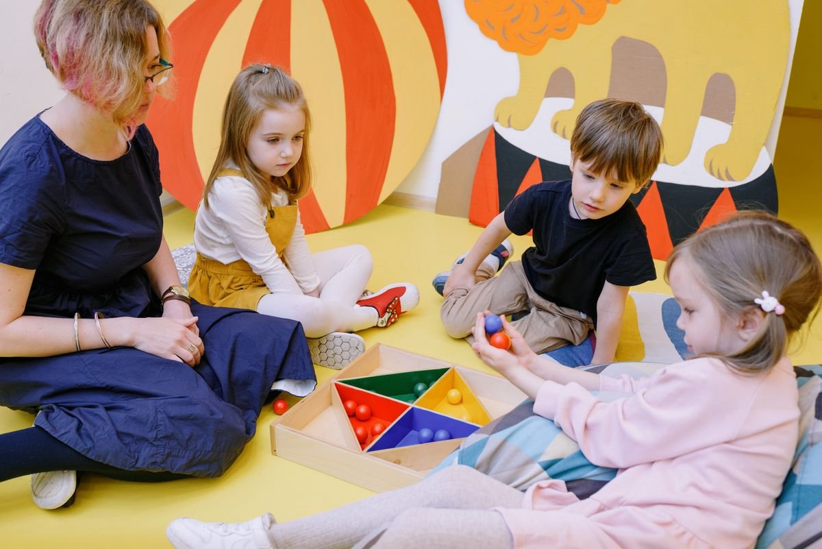 A teacher sitting on the floor with three young children around a tray of colourful shapes